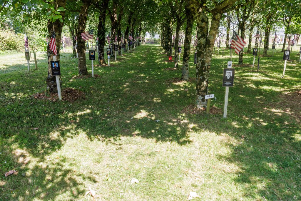 A shaded grassy lane lined with mature trees, each decorated with an American flag and a photo portrait plaque honouring a firefighter, creating a peaceful, canopy-covered memorial aisle.