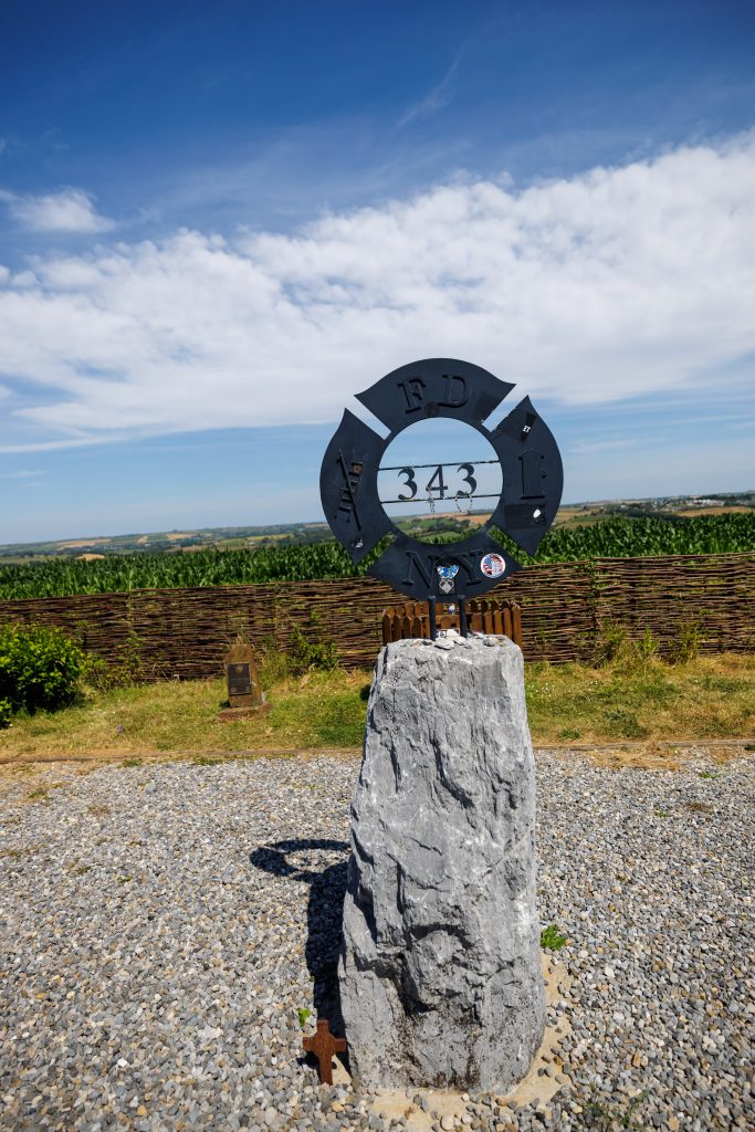 A tall, rough-cut stone marker in a gravel area featuring a black metal FDNY firefighter Maltese cross emblem mounted on top with the number 343 cut out at the center. The background shows green fields and a blue sky with light clouds.