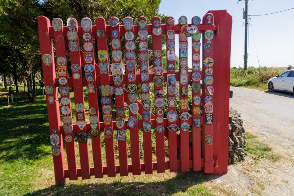 A vibrant red wooden picket gate completely covered with numerous colourful patches, badges, and insignias from various fire departments and emergency services. The gate is set outdoors on a gravel and grass pathway, flanked by green trees on the left and a road with a parked white car on the right under a bright blue sky.