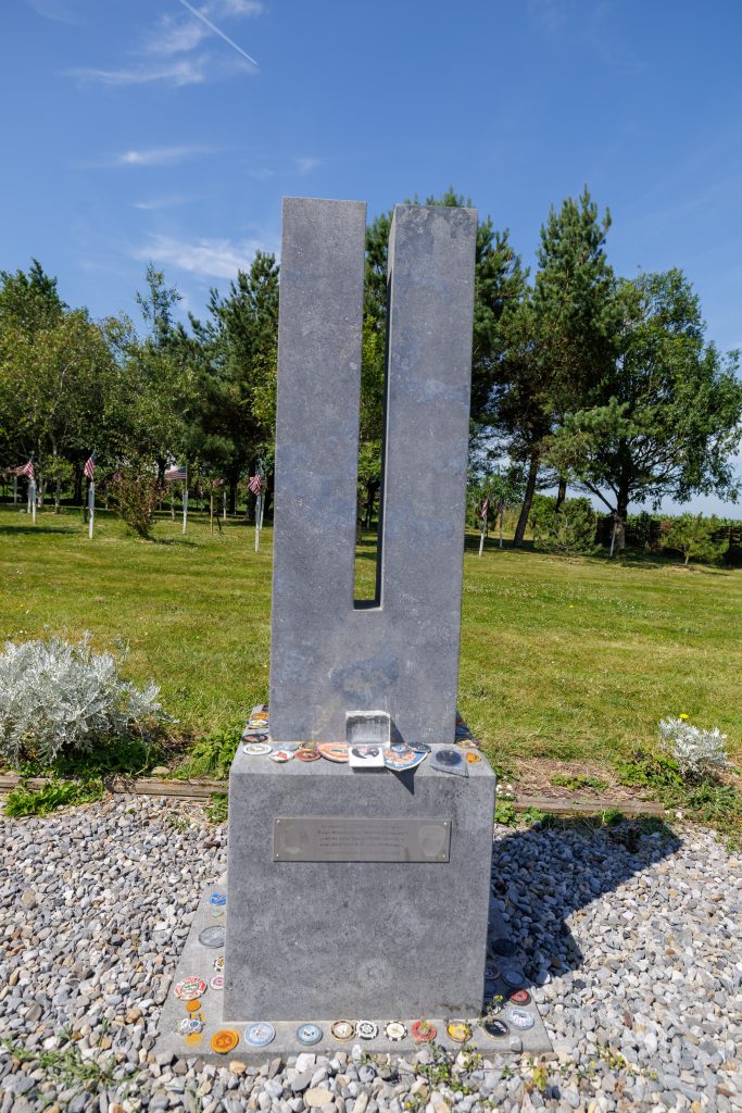 A tall, two-pillar stone memorial sculpture symbolizing the World Trade Center Twin Towers, set on a stone base covered with fire and police department challenge coins and patches. A brass memorial plaque is mounted at the base, with green grass, trees, and small American flags in the background.