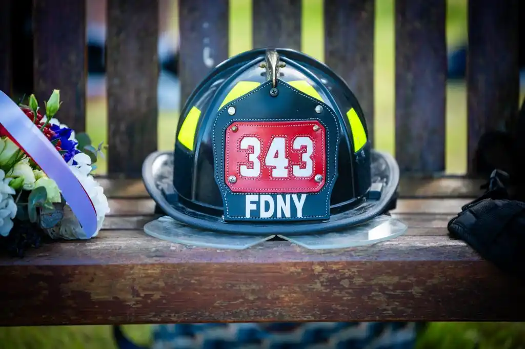 A close-up photograph of a black New York City Fire Department (FDNY) helmet resting on a slatted wooden bench. The helmet features a prominent red front shield with white numbers, framed by a black lower section and yellow reflective strips on the dome. To the left of the helmet rests a bouquet of white, red, and blue flowers wrapped in a ribbon.