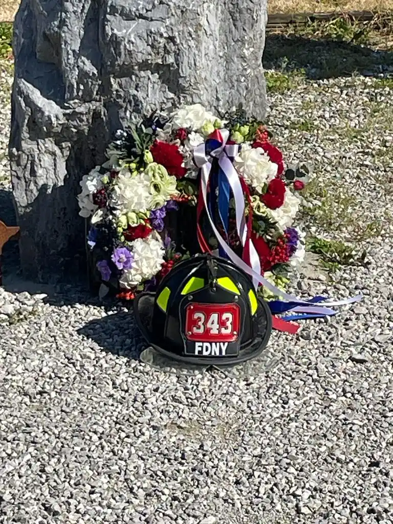 A photograph of a black FDNY fire helmet placed on a bed of grey gravel at the base of a large, rough grey stone monument. Behind the helmet rests a floral wreath composed of white, red, and purple flowers, adorned with red, white, and blue ribbons. The helmet features yellow reflective strips and a red front shield with white text.