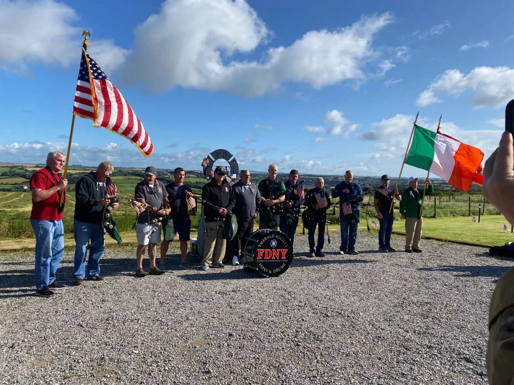A photograph of a large group of men gathered outdoors on a gravel surface in the Ringfinnan Garden of Remembrance under a bright blue sky with white clouds. Two men on the outer flanks hold large flags: the United States flag on the left and the Irish flag on the right. In the centre of the group, several men hold bagpipes. A large black bass drum rests on the ground in the middle of the assembly. The men are wearing casual clothing, including hoodies, shorts, and jeans. Rolling green fields and a distant town are visible in the background.