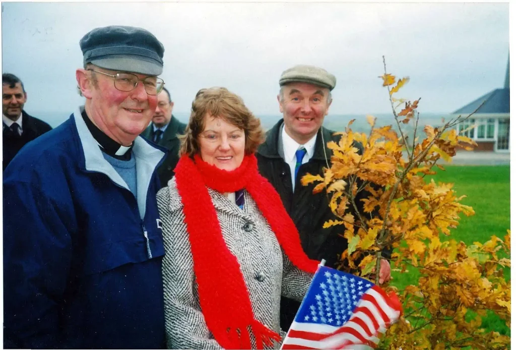 A photograph taken outdoors on an overcast day featuring four individuals. In the foreground, Kathleen Cait Murphy wearing a grey patterned coat and a bright red knitted scarf holds a small United States flag. To her left stands Father Liam O'Regan wearing a clerical collar, a light blue jumper, a dark blue jacket, and a grey flat cap. To her right, stands Virgil Horgan in a flat cap, white shirt, green tie, and dark jacket stands near a small oak tree with yellow and brown autumn leaves. A fourth man is partially visible in the background on the far left. A building and a green lawn are visible in the distant right background.