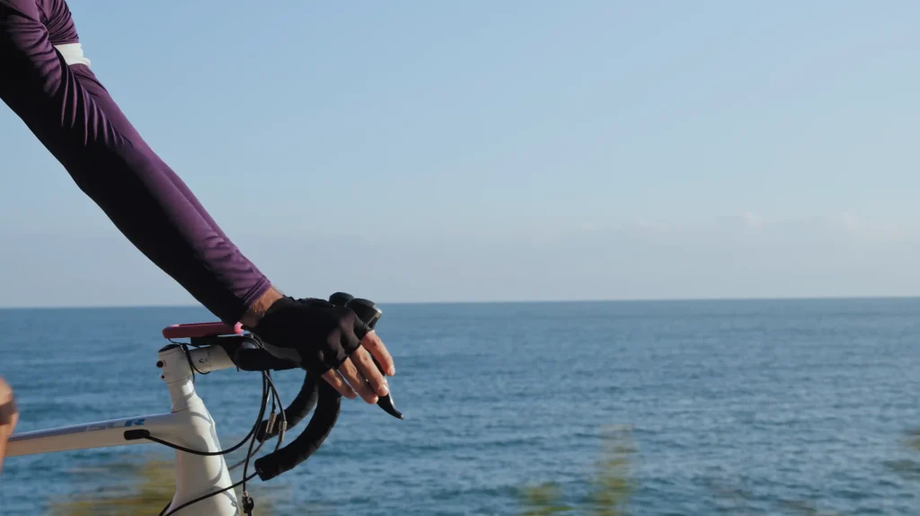 A photograph showing the arms and hands of a cyclist riding a white road bicycle. The cyclist is wearing a purple long-sleeved top and black fingerless gloves. The hands are shown gripping the drop handlebars. The background consists of a clear blue sea meeting a pale blue sky at the horizon.