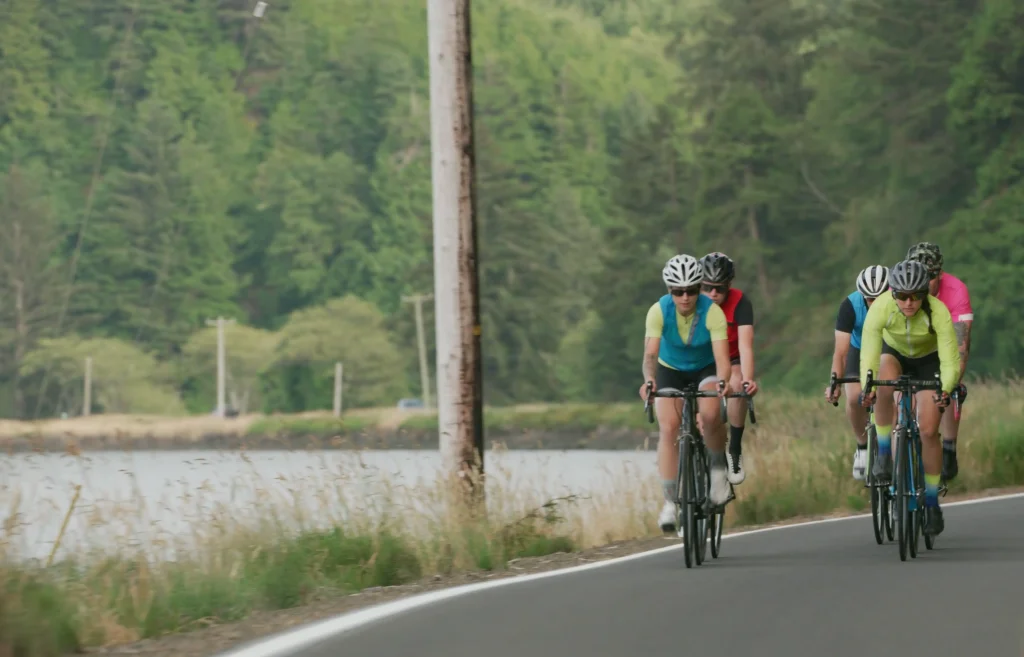 A photograph of a group of five cyclists riding in a single-file line along a paved road. They are wearing brightly coloured cycling jerseys in shades of cyan, red, yellow, and pink, along with helmets and sunglasses. The road borders a body of water on the left, with tall green trees covering a hillside in the background. A wooden utility pole stands near the edge of the road, and tall dry grass lines the shoulder.