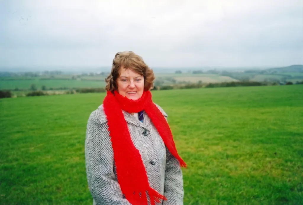 A portrait of Kathleen Murphy, founder of the Ringfinnan Garden of Remembrance in Kinsale, Ireland. She is wearing a grey patterned tweed coat and a bright red knitted scarf wrapped around her neck, standing outdoors in a lush green Irish field under an overcast sky.
