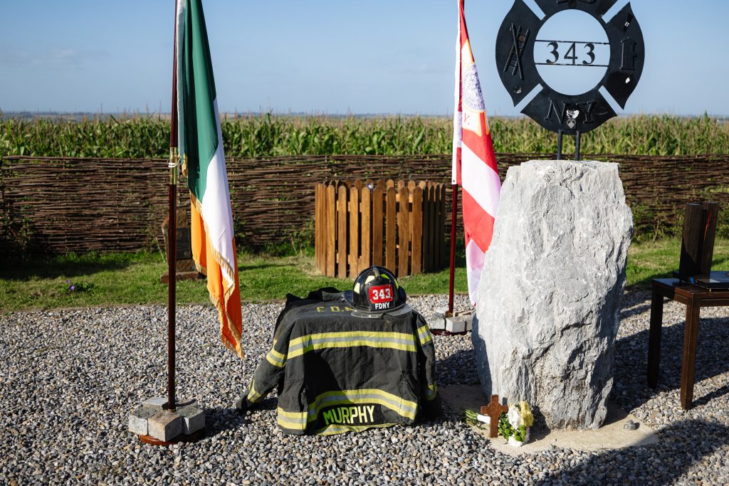 A wide, eye-level shot of the primary memorial display set on a bed of grey gravel under a bright, clear blue sky. In the centre sits a dark firefighter turnout coat topped with a black FDNY helmet bearing shield number "343". To the left stands a wooden flagpole displaying the Irish national flag (green, white, and orange). To the right stands another flagpole displaying the FDNY flag (red and white stripes with the departmental seal). Beside the flags stands a tall, upright grey limestone boulder. Mounted atop the boulder is a custom black iron cross emblem containing a firefighter ladder, fire hose, the number "343", and the letters "N Y". At the base of the stone lie flowers and a small rusted cross. In the background, a woven willow fence separates the gravel area from a green field and corn crop.