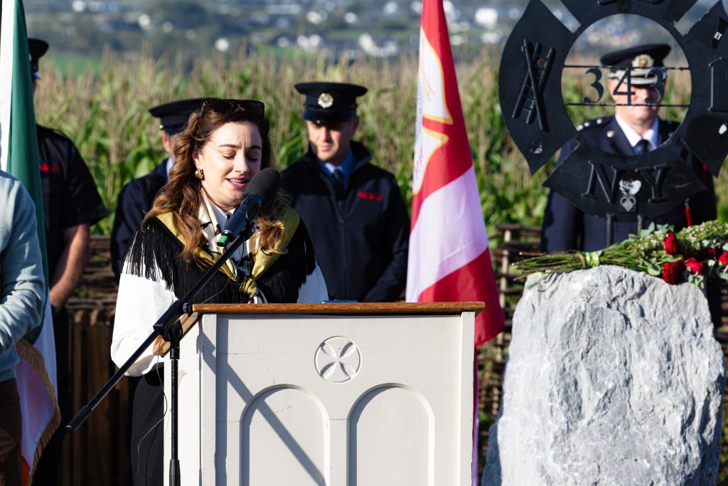 Emma Nash with long brown hair and sunglasses resting on her head speaks into a microphone at a light-coloured wooden podium featuring a Celtic cross carving. She is wearing a white top with black fringed details and a gold scarf. Behind her, a uniformed firefighter stands at attention next to an Irish flag and a dark metal FDNY 343 memorial sculpture, which sits on a large rough stone adorned with red roses. The background is filled by a field of tall maize.