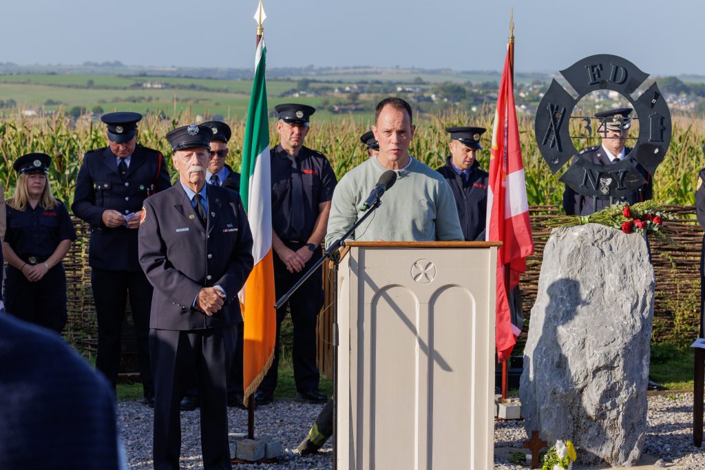 Curator of the garden, John Murphy wearing a light green jumper speaks at the wooden memorial podium. To his left stands an older man in a navy fire service dress uniform with medals, looking straight ahead respectfully. Behind them, an honour guard of firefighters in dark uniforms stands at attention in front of a field of maize. An Irish flag and the FDNY 343 memorial sculpture frame the scene, with rolling green countryside visible in the distance under a clear sky.