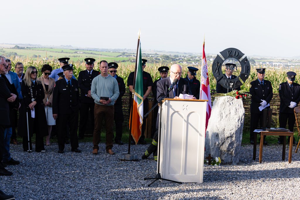 A wider shot of the 9/11 anniversary ceremony at Ringfinnan Remembrance Garden. Virgil Horgan, dressed in a dark suit and glasses reads from a piece of paper at the central podium. A gathered crowd of civilians and firefighters in dress uniforms stand in a semi-circle on the gravel, listening attentively. The memorial area features an Irish flag, a red and white flag, and the FDNY 343 sculpture. The garden is bordered by a woven wicker fence and tall crops, overlooking a scenic view of the surrounding countryside.