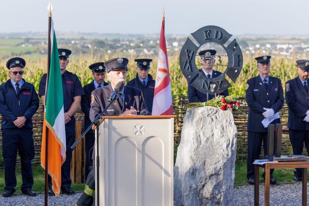 Danny Manning wearing a navy fire service dress uniform and cap, speaks into a microphone at a white wooden podium. Behind him, several uniformed firefighters stand at attention as an honour guard. The scene is framed by an Irish flag to the left and a red and white flag to the right, alongside a dark metal FDNY 343 memorial sculpture resting on a large, rough grey stone adorned with red roses. A field of tall maize and rolling green countryside fill the background.