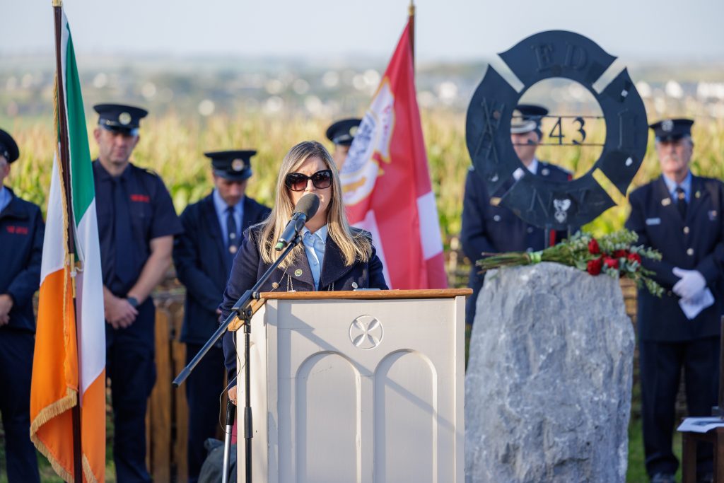 Cll Ann Bambury wearing dark sunglasses and a navy blue coat, speaks at the light-coloured wooden podium featuring a Celtic cross carving. In the background, firefighters in dark uniforms stand at attention near an Irish flag, a red and white flag, and the FDNY 343 memorial stone decorated with red roses. A field of tall crops and distant rolling hills are visible under a clear, bright sky.