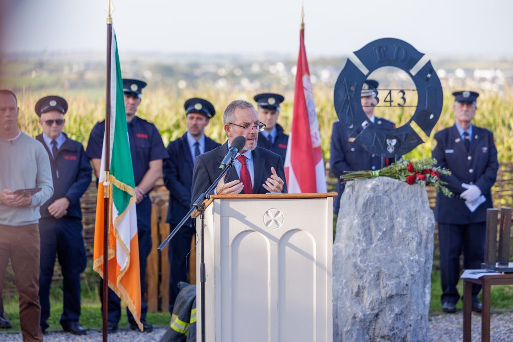 Jerry Buttimer, wearing a dark suit and a red tie, speaks at the wooden memorial podium, gesturing slightly with his hands. Behind him, an honour guard of firefighters stands respectfully in front of a woven wicker fence and a field of maize. The memorial area includes an Irish flag, a red and white flag, and the FDNY 343 sculpture on a stone base with red roses, all set against a backdrop of scenic countryside.