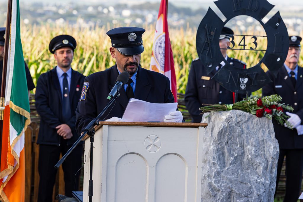 A firefighter reads from a piece of paper at a light-coloured wooden podium in the Ringfinnan Garden of Remembrance. To his left stands the Irish flag .To his right is a large rough stone bearing a dark metal FDNY 343 memorial sculpture, decorated with red roses. A floral arrangement of red, white, and blue flowers rests on the gravel nearby. An honour guard of firefighters stands in the background behind a woven wicker fence, with a field of tall maize and rolling countryside stretching into the distance.