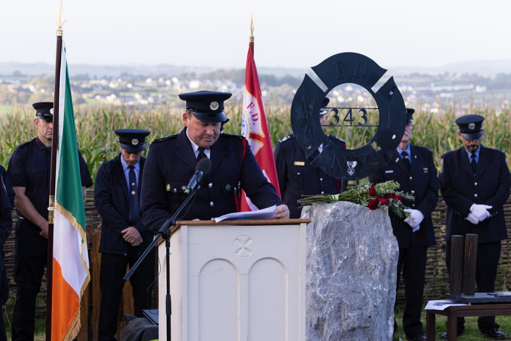 A firefighter reads from a piece of paper at a light-coloured wooden podium in the Ringfinnan Garden of Remembrance. To his left stands the Irish flag .To his right is a large rough stone bearing a dark metal FDNY 343 memorial sculpture, decorated with red roses. A floral arrangement of red, white, and blue flowers rests on the gravel nearby. An honour guard of firefighters stands in the background behind a woven wicker fence, with a field of tall maize and rolling countryside stretching into the distance.