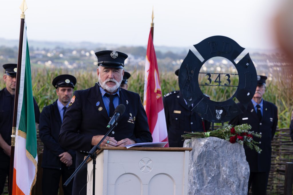 A firefighter reads from a piece of paper at a light-coloured wooden podium in the Ringfinnan Garden of Remembrance. To his left stands the Irish flag .To his right is a large rough stone bearing a dark metal FDNY 343 memorial sculpture, decorated with red roses. A floral arrangement of red, white, and blue flowers rests on the gravel nearby. An honour guard of firefighters stands in the background behind a woven wicker fence, with a field of tall maize and rolling countryside stretching into the distance.