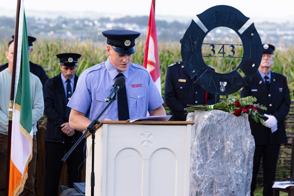 A firefighter reads from a piece of paper at a light-coloured wooden podium in the Ringfinnan Garden of Remembrance. To his left stands the Irish flag .To his right is a large rough stone bearing a dark metal FDNY 343 memorial sculpture, decorated with red roses. A floral arrangement of red, white, and blue flowers rests on the gravel nearby. An honour guard of firefighters stands in the background behind a woven wicker fence, with a field of tall maize and rolling countryside stretching into the distance.