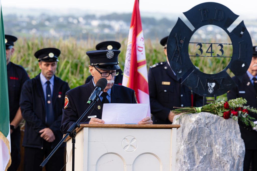 Danny Manning reads from a piece of paper at a light-coloured wooden podium in the Ringfinnan Garden of Remembrance. To his left stands the Irish flag .To his right is a large rough stone bearing a dark metal FDNY 343 memorial sculpture, decorated with red roses. A floral arrangement of red, white, and blue flowers rests on the gravel nearby. An honour guard of firefighters stands in the background behind a woven wicker fence, with a field of tall maize and rolling countryside stretching into the distance.