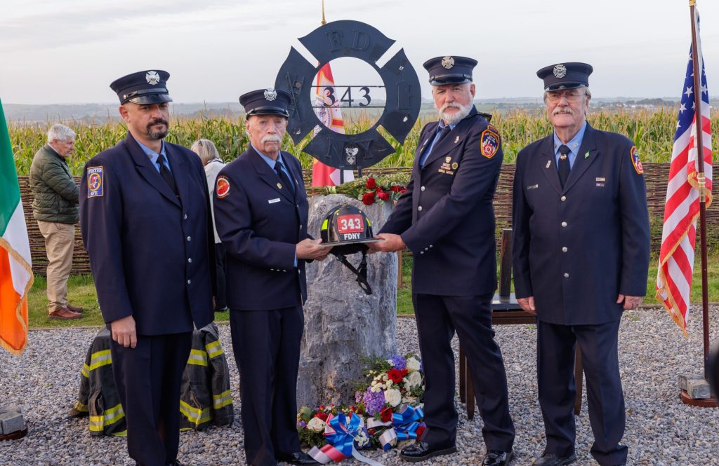 Danny Manning holds a memorial firefighter helmet with 3 other uniformed firefighters in front of the 343 Maltese Cross Memorial in Ringfinnan Garden of Remembrance
