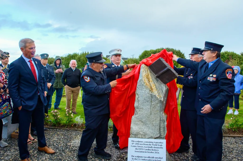 U.S. Ambassador Edward Walsh smiling as he watches a group of retired FDNY firefighters in dress uniforms pull back a bright red cloth to unveil a new stone monument topped with a piece of World Trade Center steel.