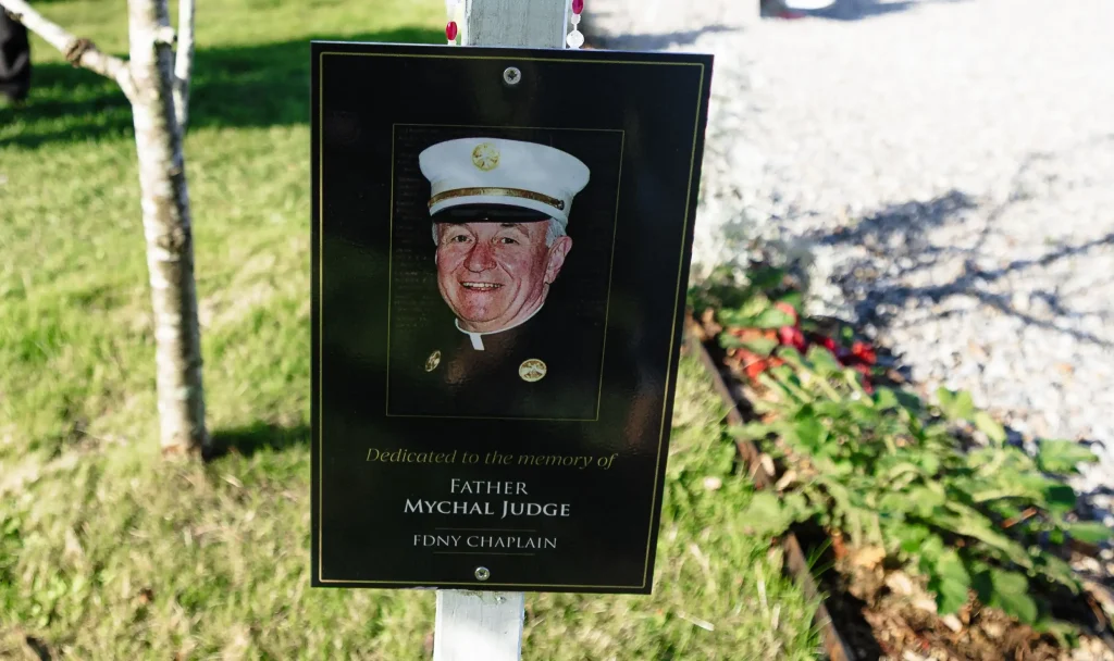 Closeup photo of the plaque of Chaplain Mychal Judge screwed to a white post in the Ringfinnan Garden of Remembrance. Fr Judge is wearing is FDNY uniform.