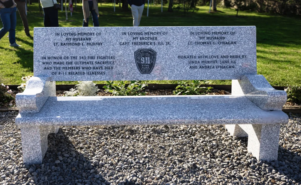 Polished grey granite memorial bench situated on gravel in the Ringfinnan Garden of Remembrance, engraved with dedications to Lt. Raymond E. Murphy, Capt. Frederick J. Ill, Jr., and Lt. Thomas G. O'Hagan, and honouring the 343 firefighters lost on 9/11.
