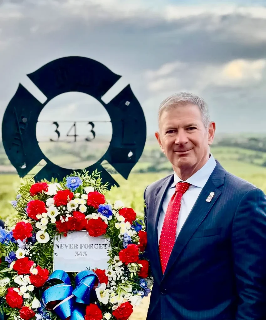 The US Ambassador to Ireland, Edward S Walsh stands beside the Maltese Cross Memorial in the Ringfinnan Garden of Remembrance with a flower tribute and sign "never forget 343" to mark the 24th anniversary of 9/11.