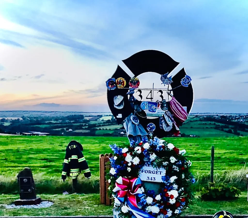 Maltese Cross Memorial in the Rinfinnan Garden of Remembrance with floral wreaths placed at the base and hanging of the front. Fire Department memorabila cover the circular top section of the memorial. There is a FDNY jacket hangng off a fence post behind. The sky highlights that the picture was taken in the evening of a sunny day.